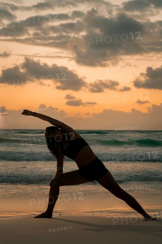 A woman performs a yoga pose on a serene beach at sunrise, surrounded by gentle waves and a vibrant sky. The scene captures tranquility and focus in a natural setting of Tulum, Mexico