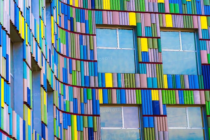 A close-up view of a modern building's facade featuring a vibrant, multicolor pattern with distinct blue, yellow, and green hues and curved elements around windows