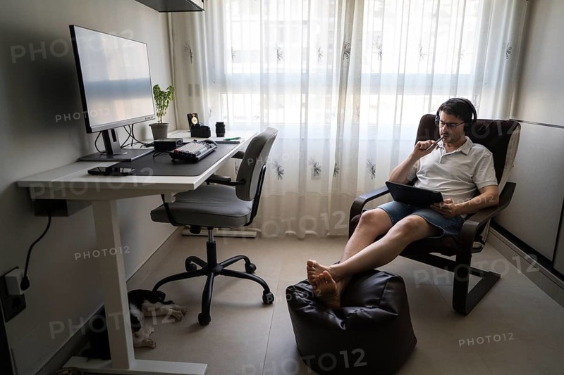 A man comfortably seated at home, wearing headphones and using a tablet to study Mandarin online. The room is bright, with a desk and computer nearby