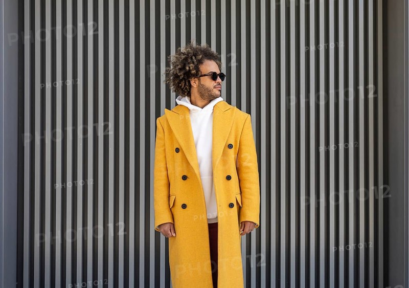 A stylish afro man in a mustard yellow coat stands confidently in front of a modern striped background, exuding casual elegance while wearing sunglasses