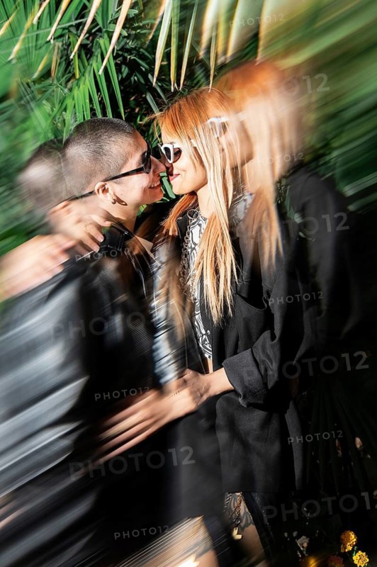 A stylish lesbian couple shares a tender kiss, surrounded by vibrant greenery. The atmosphere is filled with love and intimacy, captured in a dynamic and colorful scene