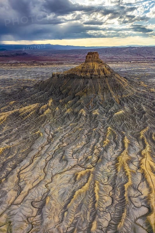 A striking aerial photograph captures the ethereal landscape of Factory Butte during sunset in Utah, highlighting the intricate textures and formations of the terrain. The play of light accentuates the golden hues against the rugged earth