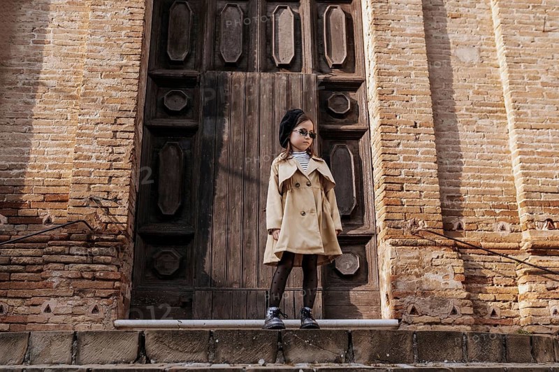 A stylish young girl in a trench coat and sunglasses stands confidently by a historic wooden doorway in Civitanova, Italy. The brick architecture adds to the picturesque scene