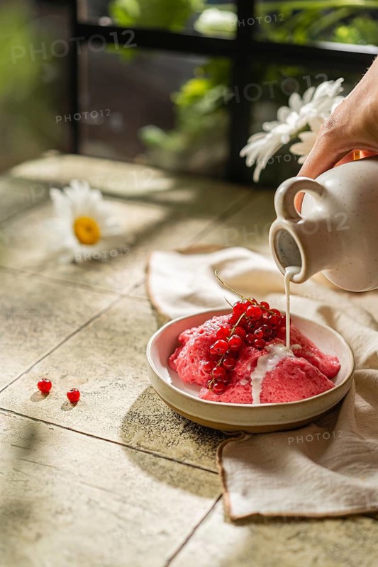 Cropped unrecognizable person hand's pouring cream over homemade berry mousse garnished with fresh currants, served in a rustic ceramic bowl