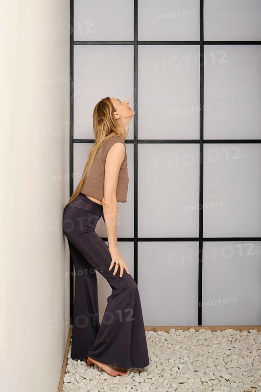 A woman stands in a yoga pose against a wall in a room with a grid design. She wears comfortable clothes and focuses on her balance. The floor has stones, creating a natural look in the space