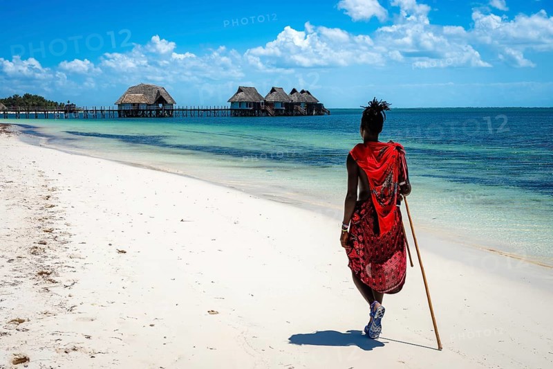 A Maasai man in traditional red attire walks away from the camera along a pristine white sand beach in Zanzibar, Tanzania