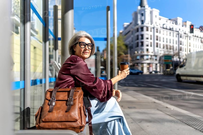 Senior woman with gray hair and glasses sitting at a city bus stop on a sunny day, holding a disposable coffee cup and waiting for public transportation with her backpack beside her
