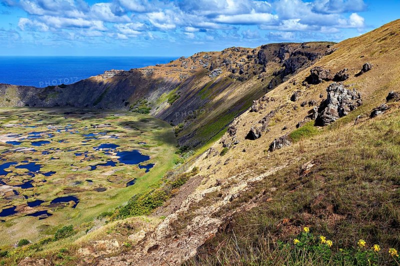 Rocky volcanic slopes overlooking a crater with water and the ocean behind it, the Ranu Kau volcano on Easter Island in the Pacific Ocean
