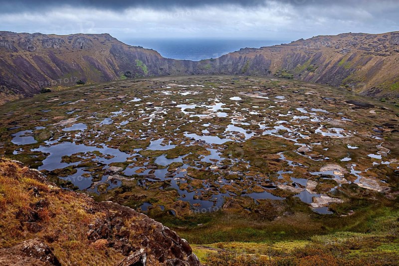 Large crater with expanses of water surrounded by rocky and grassy walls under cloudy skies, the Ranu Kau volcano on Easter Island in the Pacific Ocean