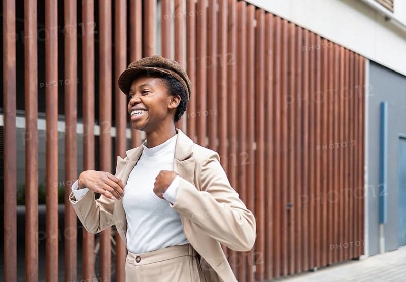 A black woman in a beige outfit and cap walks confidently in an urban environment The setting features wooden paneling, reflecting a stylish city vibe