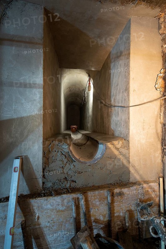 A partially constructed underground tunnel with visible concrete supports and unfinished surfaces. Electrical wiring and construction tools are visible, showcasing work in progress