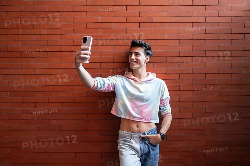 A young man smiles while taking a selfie with his smartphone against a vibrant red brick wall. He is wearing a colorful cropped hoodie and jeans, creating a casual vibe