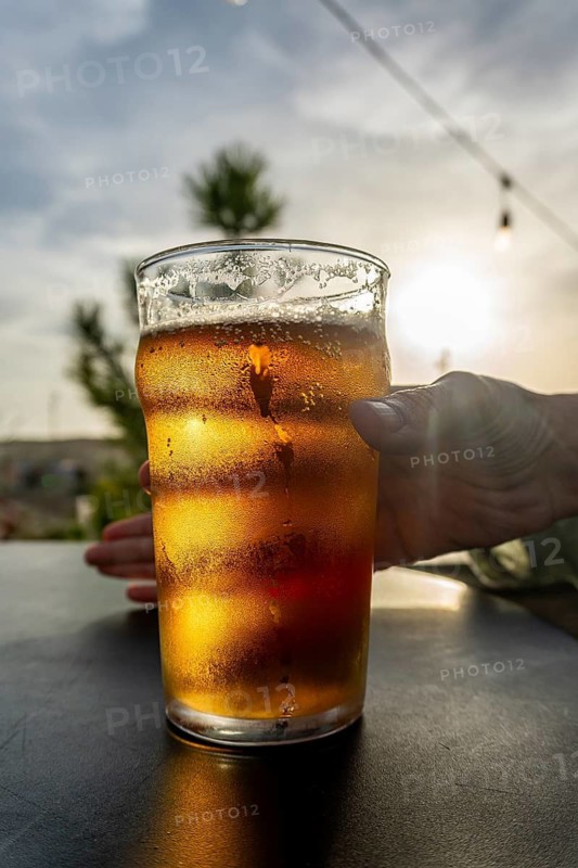 Hand holding condensation covered pint of cold beer on a table at sunset, golden light and foamy bubbles evoke summer holiday relaxation on a terrace by the coast
