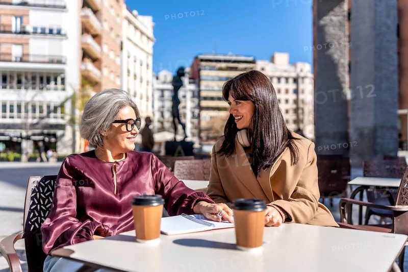 Two women of different generations enjoying coffee and a lively conversation at an outdoor cafe on a sunny day, collaborating on an open notebook together