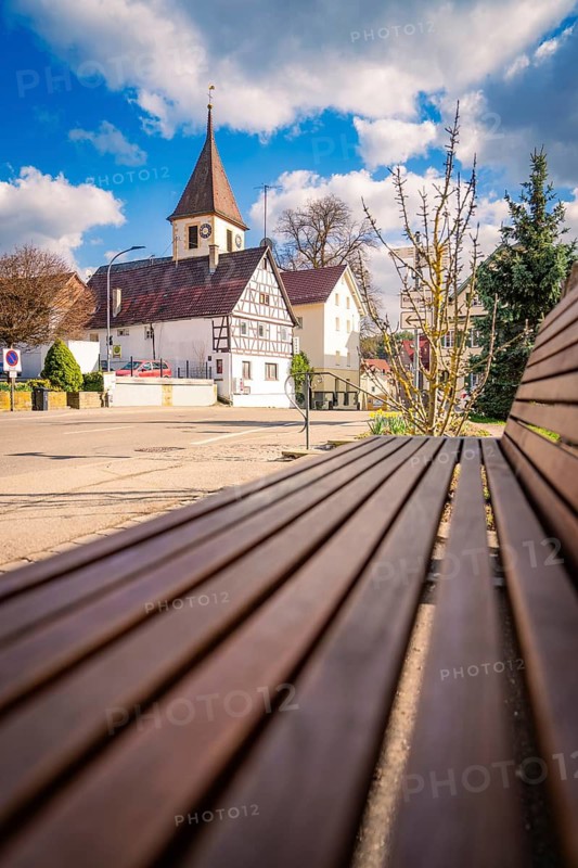 Wooden bench in a quiet suburb, with half-timbered church and houses in the background under cloudy sky, Deufringen, Aidlingen, Böblingen district, Germany