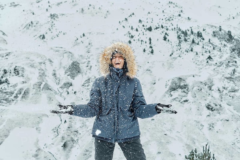 A joyful person stands in falling snow, clad in a winter jacket and gloves, at Astun ski resort in the Huesca Pyrenees. The snowy landscape creates a serene backdrop