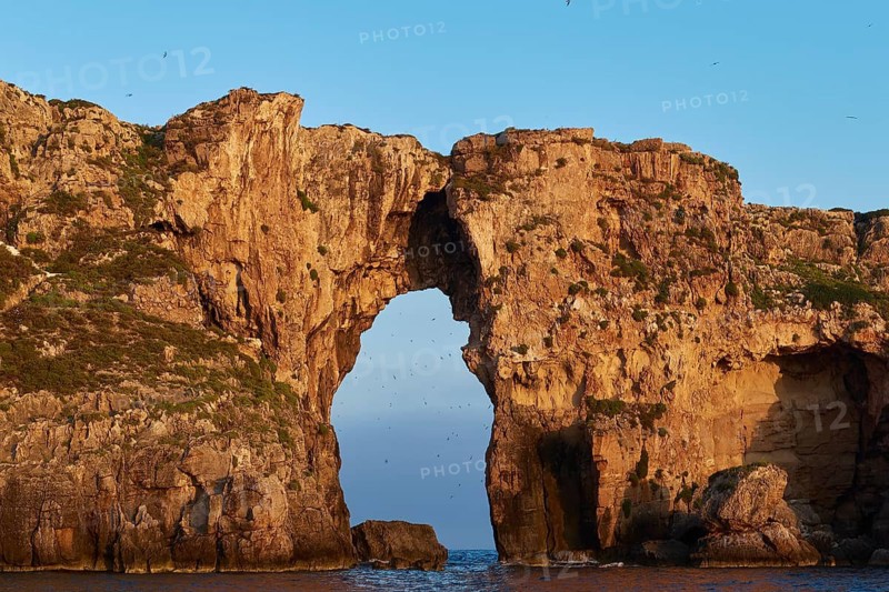 An imposing rock gate stretches across the calm sea under clear skies, morning light, Tsichli Babas islet, Sfaktiria island, Navarino Bay, Pylos, Peloponnese, Greece