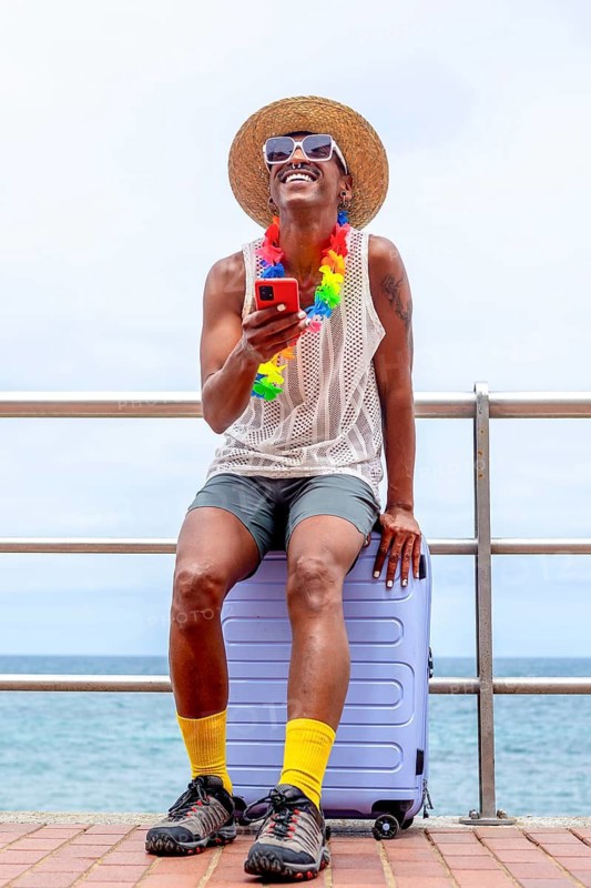 An LGBTQIA+ man dressed in vibrant attire, seated on a suitcase by the sea, joyfully preparing for holiday adventures, embodies pride and anticipation