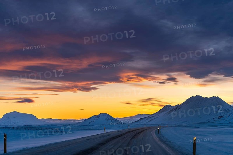 A serene winter drive through Iceland's snowy landscape at dusk. The vibrant sunset colors the sky, contrasting with the pristine, snow-covered mountains and road