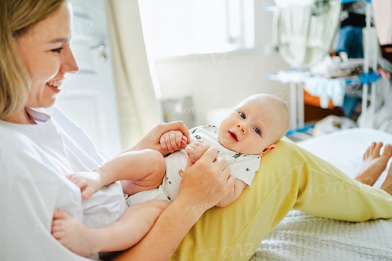 A smiling mother holds her baby on her lap at home, enjoying precious bonding moments. The bright room adds warmth, emphasizing maternal love and connection