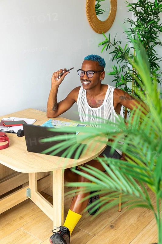 LGBTQIA+ man excitedly planning a holiday trip on a laptop. Surrounded by a cozy setting with plants, they exude joy and anticipation. Embracing diversity and adventure
