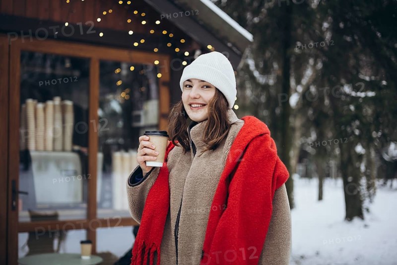 A cheerful young woman, standing outside a cozy cafe, enjoys a warm beverage on a cold winter day. She's dressed in a stylish red scarf and a white beanie, with twinkling lights and snow-covered trees in the background