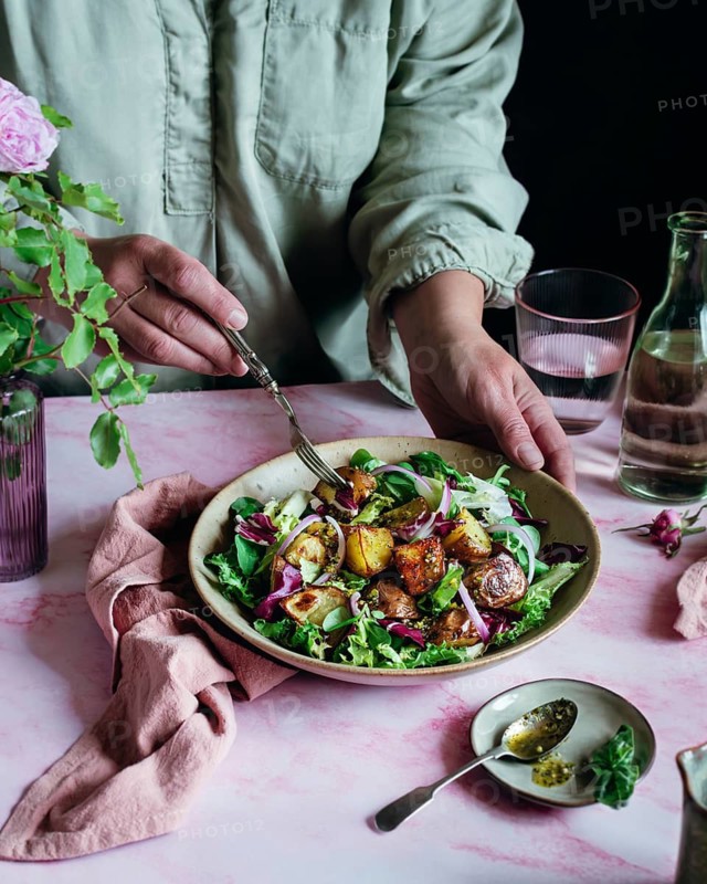 A person in a light shirt prepares a vibrant potato salad with fresh greens, roasted potatoes, and red onion on a pink table. A fork is held, ready to toss the salad