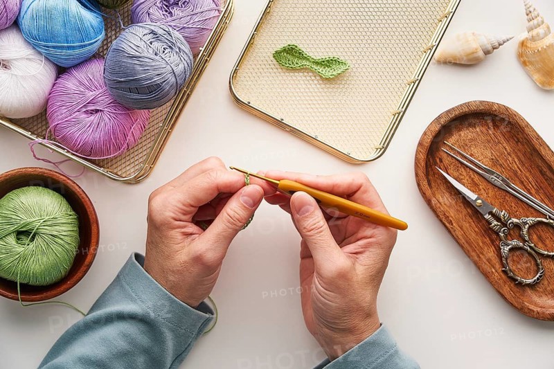 Top view cropped unrecognizable hands of a woman crocheting in her studio. Surrounded by colorful yarns and crafting tools, creating detailed handmade designs
