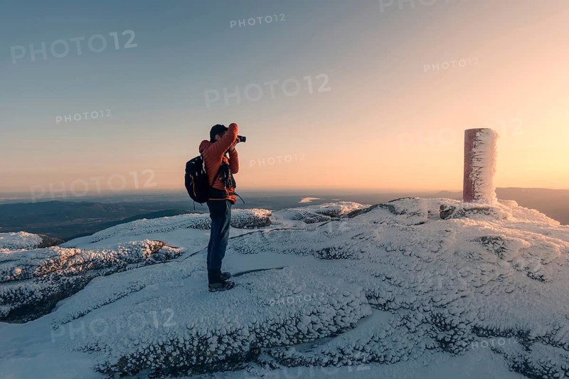A photographer stands on the snowy summit of Siete Picos, capturing the serene sunrise over the Guadarrama Mountains, Madrid. The scene is peaceful and breathtaking
