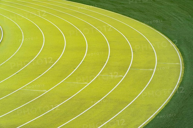 Overhead view of vibrant green lanes vividly marking the curves on an outdoor athletics stadium, capturing the precise lines and lush greenery beside the track