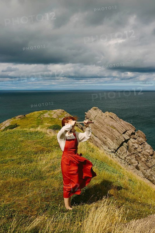 A woman in a vintage red dress and sunglasses using a spyglass on a windy coastal cliff at Middle Cove, Newfoundland & Labrador, Canada. The dramatic clouds and ocean view enhance the scene's adventurous feel