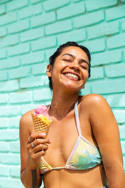 A cheerful latin woman in a pastel bikini enjoys a refreshing ice cream cone on a bright summer day. She smiles blissfully against a vibrant turquoise brick wall