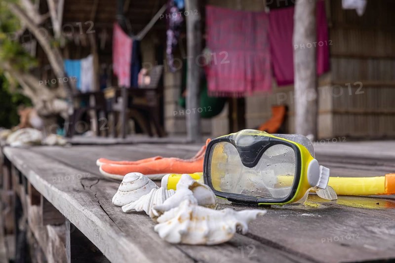 Snorkeling gear with seashells are laid out on a rustic wooden table on Kri Island, with blurred background showing a traditional homestay and laundry hanging, in the Raja Ampat archipelago, Indonesia