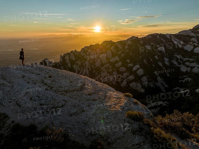 A lone figure stands on a rocky cliff at Montserrat, Catalonia, as the sun rises, casting warm light over the rugged landscape and distant hills