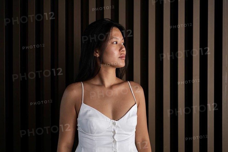 A young Chinese woman in a stylish white dress stands confidently against a wooden backdrop. The warm summer ambiance creates a serene city vibe as she gazes thoughtfully