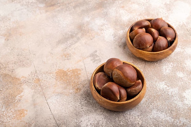 Wooden bowl with raw edible ?hestnuts on brown concrete background, side view, copy space, minimalism
