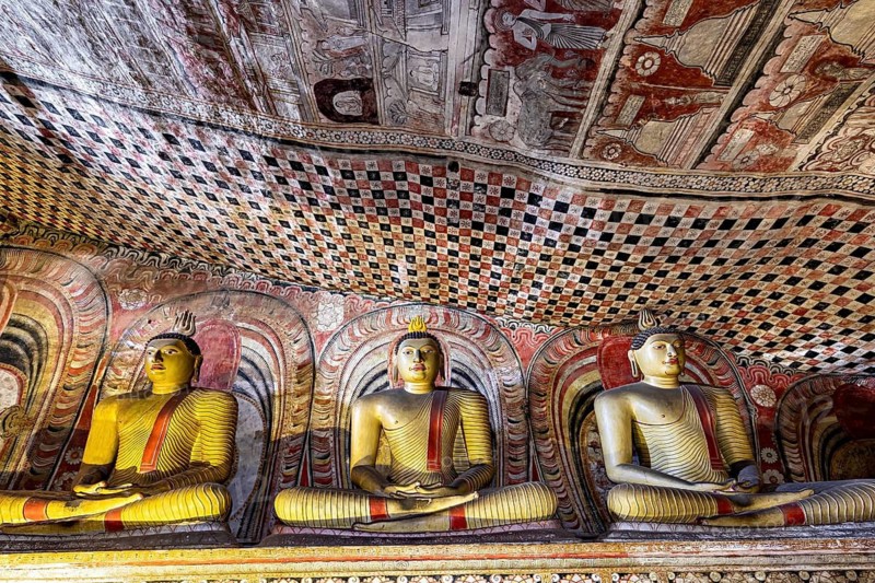 Three Buddha statues under artfully painted vaults in deep colors, The cave temple of Dambulla in Sri Lanka