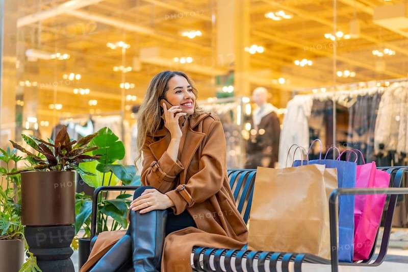 Young woman sits on a bench in a modern mall, smiling and talking on her smartphone with shopping bags beside her during a relaxed, stylish shopping break