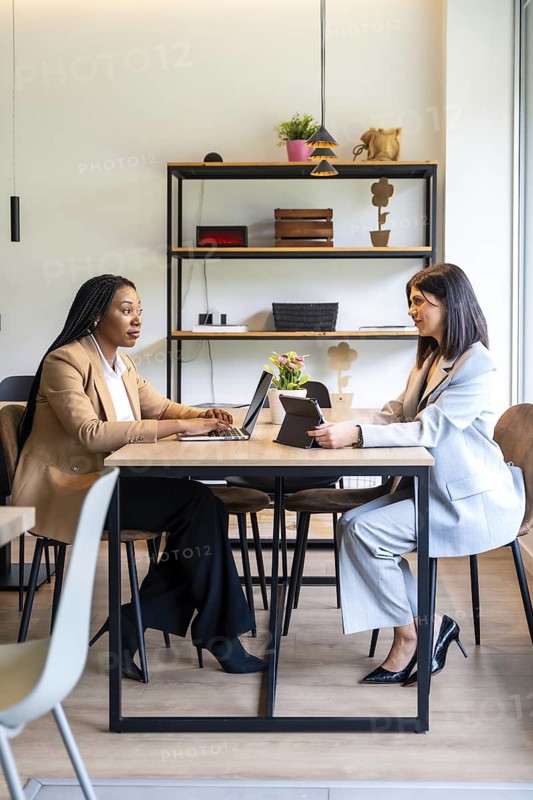 Two businesswomen are working together on their laptops and tablets in a hotel lobby, discussing business strategies