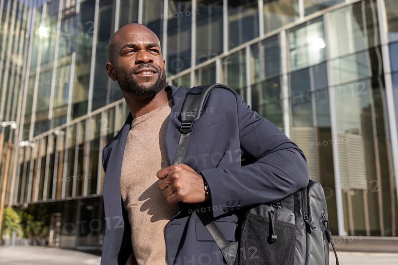 A confident cuban man in a blazer and sweater, carrying a backpack, stands outside a modern office building. The sun shines on the glass facade, casting a warm glow