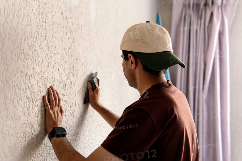 An unrecognizable man wearing a baseball cap is seen sanding a textured wall, part of his home remodeling project. Focus on hand and tool usage for precise DIY work