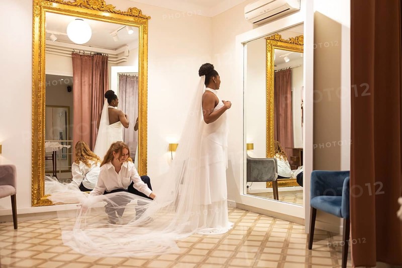 Bride stands elegantly in a fitting room, her long veil being adjusted by an assistant. The room features large mirrors and a gold framed wall mirror, creating a sophisticated atmosphere