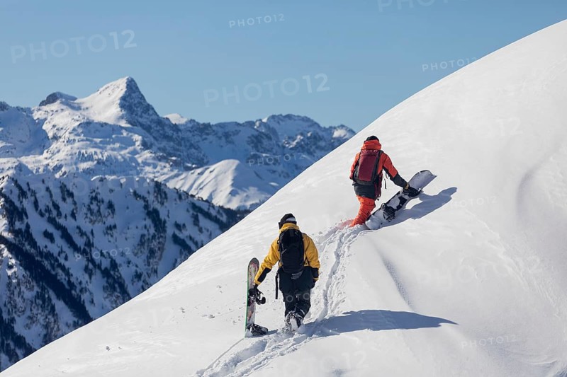 Two snowboarder friends trek a snowy mountain slope under clear blue skies, with majestic peaks in the background, capturing the thrill and beauty of winter sports and exploration