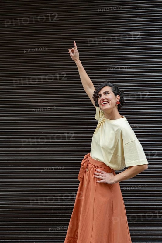 A fashion woman in a beige shirt and orange skirt smiles and points upwards against a textured background, conveying positivity and aspiration in a candid pose