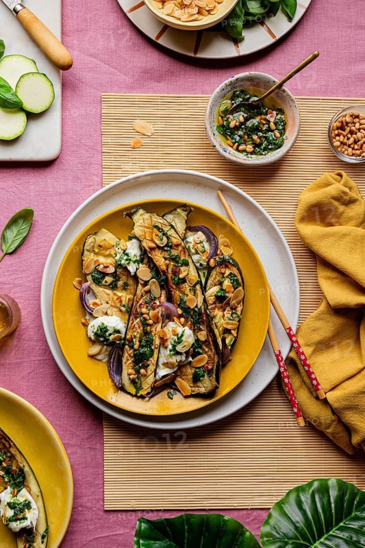Top view of gourmet roasted courgettes topped with mozzarella, fresh basil, and toasted almonds, served on a yellow plate with a vibrant pink background