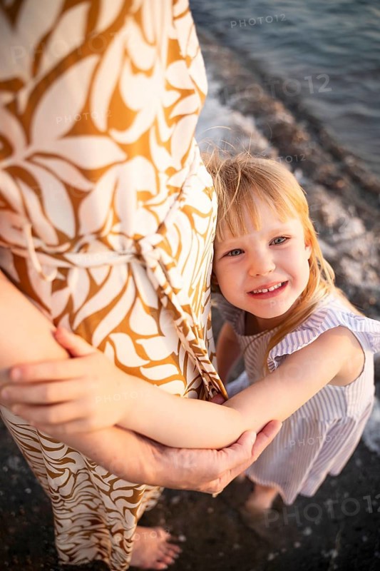 A happy little girl hugs her mother tightly while standing barefoot on a beach in Italy. She looks up with a joyful smile, capturing a warm and loving moment by the shore at sunset