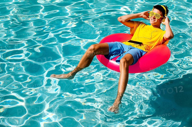 A young teenage boy in a yellow t-shirt and blue shorts relaxes on a red pool float while listening to music through headphones. The sunlit water sparkles around him, depicting summer leisure