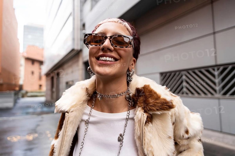 A fashionable woman with sunglasses smiles with tooth gem confidently looking at camera while wearing a fur coat and jewelry. The city street setting enhances her modern style and bold expression