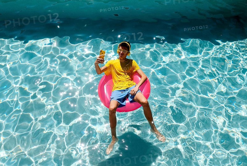 A teenage boy in a yellow shirt and shorts lounges on a pink float in a sparkling blue pool. They are wearing headphones and sunglasses, holding a phone for a selfie, enjoying a sunny day