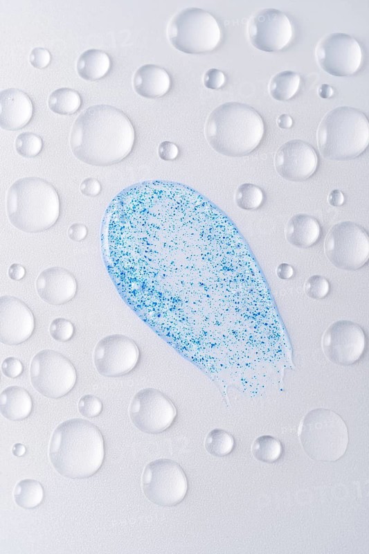 Close-up of face scrub smears with blue micro-exfoliating particles, surrounded by water droplets on a light background Perfect for skincare and beauty themes